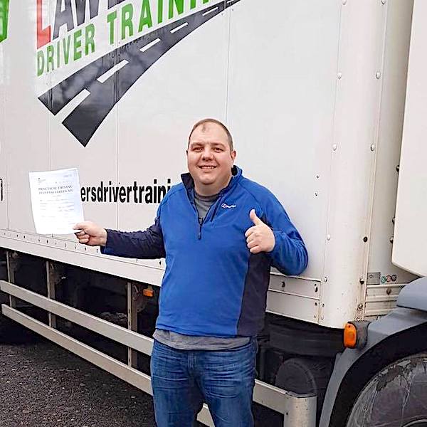 Newly qualified HGV Class 2 category C driver with thumbs up holding pass certificate in front of HGV truck