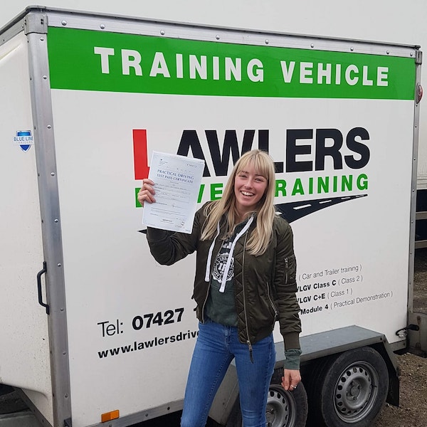 Smiling newly qualified lady trailer towing driver holding b+e pass certificate in front of  trailer training vehicle