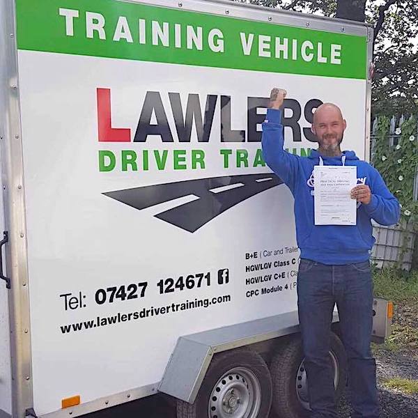 Newly qualified trailer towing driver with arm aloft holding b+e pass certificate in front of  trailer training vehicle